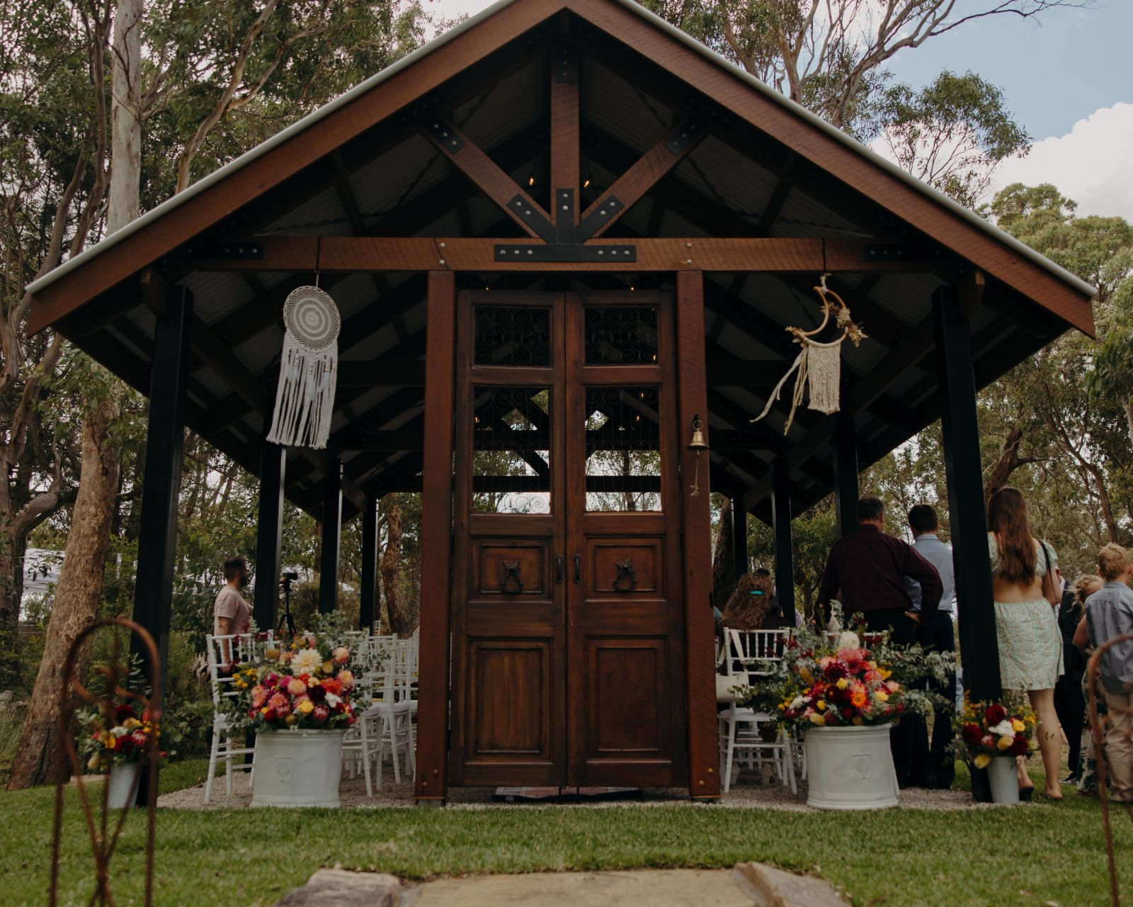 A wedding ceremony under the timber chapel at Yarrabee, urns of native and garden flowers either side of the doors